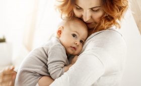 Beautiful young mother tenderly hugs her little cute child. Mom looking at her kid and smiling while baby looks aside and snuggling to mom chest. Happy family moments