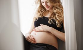 Close up detail of young attractive blonde pregnant mother in black outfit sitting on window sill in bedroom, touching and looking at belly with happy expression. Maternity concept.