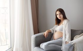 Husband taking picture of his charming pregnant wife sitting near window on armchair, smiling with cute and tender expression at camera, being calm and happy while waiting for daughter to be born.