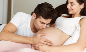 Smiling young pregnant wife lying in bed with her husband in the bedroom, man is kissing her belly