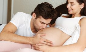 Smiling young pregnant wife lying in bed with her husband in the bedroom, man is kissing her belly